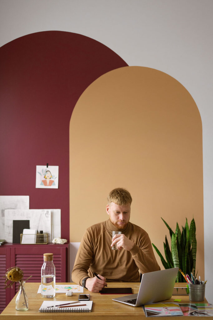 An adult in their mid-30s sits on a wooden desk using a digital pen tool and a laptop. Behind him is a wall with two colored arches painted over one another to create an interesting space on an otherwise white wall. This is a great  budget-friendly creative home decor strategy. The colors are burgundy and tan. There is also a burgundy piece of furniture in front of the burgundy painted arch. The space feels both inspiring and creative as well as professional.