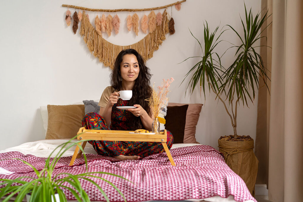 A young native woman sits on her bed wearing traditional textiles in her clothing in the form of overalls while a handmade piece of art is hung behind her. Using items belonging to your cultural heritage is a great way to make budget-friendly creative home decor that is personal.