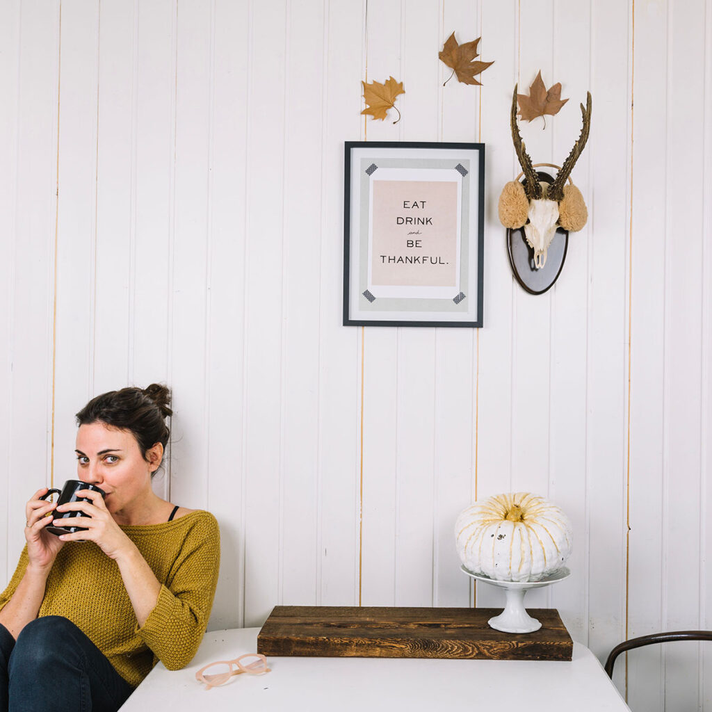 A young woman sits at a white table with her pink glasses on the table. The table has a piece of dark wood and two found objects: a fake pumpkin on a bowl stand. Above the table there are three leaves pinned in a sort of arch above two focal wall decor items. One of the items looks like a homemade poster using decorative tape in a glass matted frame. Next to it is a mounted steer head that looks like it was thrifted. The steer head is wearing tan ear muffs. This a great example of creating a  budget-friendly creative home by pairing interesting things as wall decor.