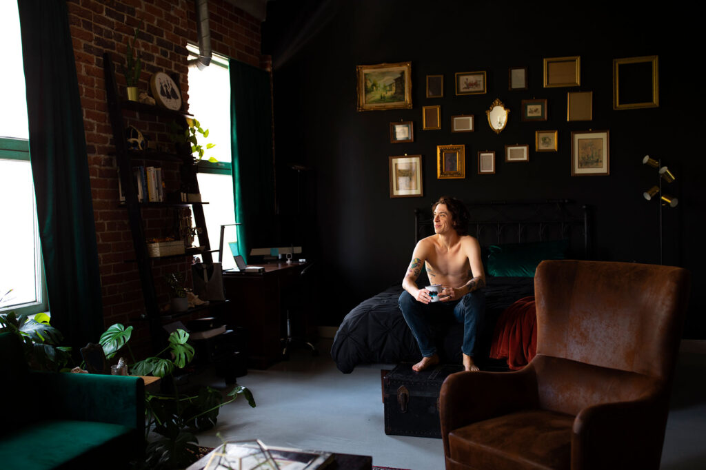 Young adult at home sitting on bed in a room with dark moody walls and a creative photo gallery behind him. The adult's aesthetic seems gothic with accents of baroque decor in the form of dark velvet colored accents and furniture. 